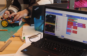 coding displayed on a laptop while students hands connect it to robotic vehicle