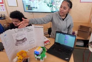 Student points to outline on a display board with care bot and laptop up front